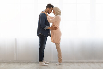 Wall Mural - Romantic muslim spouses expecting baby standing together against window at home