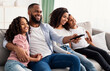 © Prostock-studio - Happy young black family watching television sitting on sofa