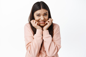 Awkward young woman feel uncomfortable, squeeze teeth and looking guilty, feeling worried, standing anxious or nervous against white background, wearing hoodie