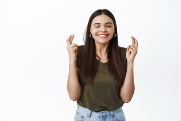 Wall Mural - Image of smiling hopeful girl think positive, making wish with closed eyes and big grin, cross fingers for good luck, standing over white background