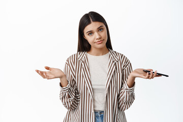 Wall Mural - Dont know. Clueless young woman have nothing to say, holding smartphone and shrugging shoulders confused, standing against white background