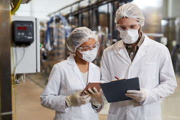 Waist up portrait of two workers wearing masks and lab coats while discussing production at chemical plant, copy space