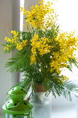 Wall Mural - a bouquet of yellow mimosa in a glass vase and a green lantern by the window