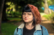 © Arturo - Portrait of a young caucasian woman with black and red hairstyle, jean jacket and vintage glasses looking at camera with serious expression. Green trees and park in the background