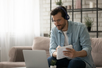 Confident male student in wireless headset sit on sofa watch education video online from university web platform write take notes. Young man working from home improve skills on distant lecture webinar