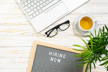 Hiring Now banner with text on the letterboard, flower in a pot, lap top and cup of herbal tea on white wooden background. Concept of hiring and recruitment.