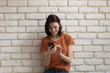 Youth and tech. Happy smiling millennial female in casual clothes engaged in using smartphone app texting messaging. Laughing young woman stand near white brick wall having fun with cell. Copy space