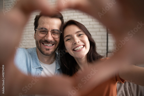 Happy Valentine day. Selfie portrait of romantic family couple holding heart of joined fingers looking at camera. Happy young spouses feel thankful grateful to destiny for meeting love strong marriage