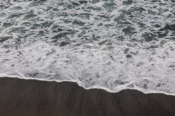 Wall Mural - Waterscape background. Black sand beach with white milky foam waves. Nature and environment concept. Black and white contrast. Daylight. Copy space. Volcanic beach in Bali