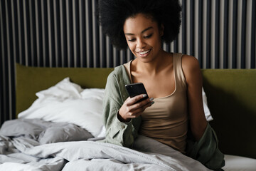 Wall Mural - Happy african american girl using mobile phone while sitting in bed