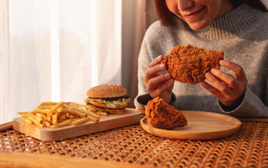 Wall Mural - Closeup image of a young asian woman holding and eating fried chicken with hamburger and french fries on the table at home