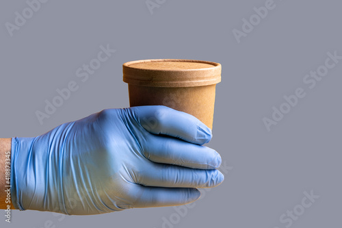A man\'s hand in a rubber glove holds out a paper disposable cup with a drink or soup on a gray background.