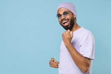 Wall Mural - Young smiling happy cool unshaven black dark-skinned african man 20s in violet t-shirt purple hat glasses do winner gesture clench fist celebrating isolated on pastel blue background studio portrait.