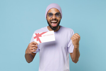 Wall Mural - Young happy unshaven student black african man in violet t-shirt hat glasses hold gift voucher flyer mock up do winner gesture clench fist clebrate isolated on pastel blue background studio portrait.