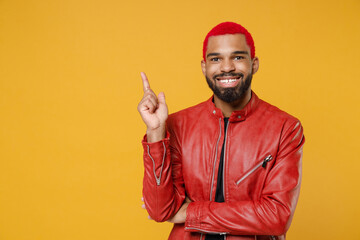 Poster - Young african dark-skinned smiling man 20s with funky trendy pink hairdo in red leather jacket pointing index finger aside on workspace area isolated on yellow orange color background studio portrait.