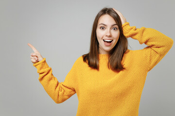 Wall Mural - Young excited happy surprised shocked caucasian woman in casual knitted yellow sweater point index finger aside on copy space area mock up holding head isolated on grey background studio portrait