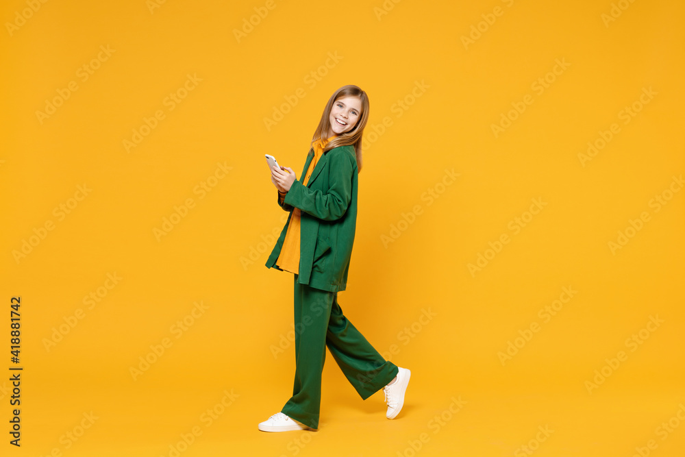 Full length side profile view of little blonde smiling kid girl 12-13 years old wearing casual clothes hold mobile cell phone walk isolated on yellow background children Childhood lifestyle concept. - obrazy, fototapety, plakaty 