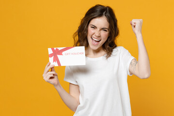 Young happy overjoyed fun caucasian woman in white basic t-shirt point index finger on gift voucher flyer mock up do winner gesture clench fist celebrate isolated on yellow background studio portrait.