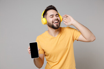 Wall Mural - Young caucasian bearded happy student man 20s in yellow basic t-shirt with closed eyes listen to music in headphones enjoying hold mobile cell phone isolated on grey color background studio portrait