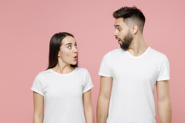 Wall Mural - Young shocked fun couple two friends bearded man brunette woman in white basic blank print design t-shirts posing looks to each other say wow isolated on pastel pink color background studio portrait