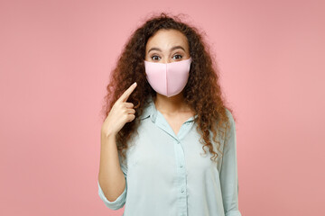 Wall Mural - Young black african curly woman in blue shirt point index finger on face mask to safe from coronavirus virus covid-19 during pandemic quarantine isolated on pastel pink background studio portrait