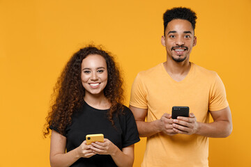 Wall Mural - Young couple two friends together family african man woman 20s in black t-shirt holding using mobile cell phone chatting in social network dating isolated on yellow color background studio portrait.
