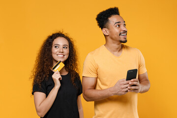 Wall Mural - Young couple two friends together family african dreamful man woman 20s in black t-shirt holding mobile phone credit bank card shop online look aside isolated on yellow background studio portrait.