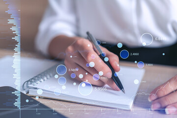 A woman trader in formal wear writing down some ideas to research stock market to proceed right investment solutions. Wealth management concept. Hologram Forex chart over close up shot.