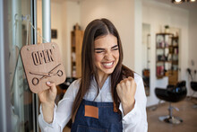 Young Woman Opening A Beauty Store And Looking Very Happy - Small Business Concepts. Smiling Owner Of Hair Salon Standing With Sign Open And Leaning On Glass Door