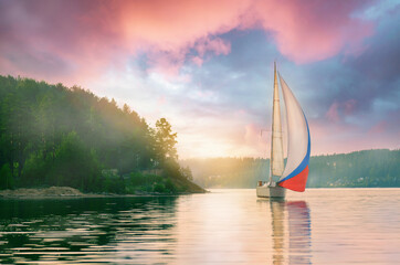 Wall Mural - Sailing yacht sailing under a beautiful sunset, reflection of the sparkling sun in the water on the shore light fog.