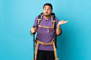 Young caucasian man with backpack and trekking poles isolated on blue background making doubts gesture