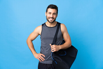 Young sport man with sport bag isolated on blue background posing with arms at hip and smiling