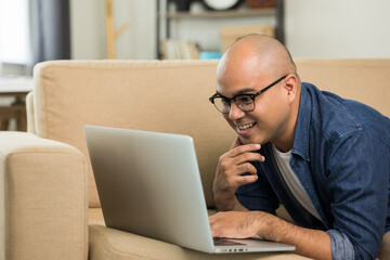 Indian man using laptop on sofa in living room texting on laptop sending message or chatting with online social media.