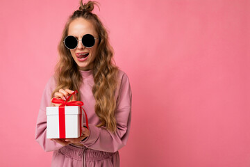 Shot of beautiful surprised positive young blonde curly woman isolated over pink background wall wearing pink sport clothes and sunglasses holding gift box and unboxing surprise