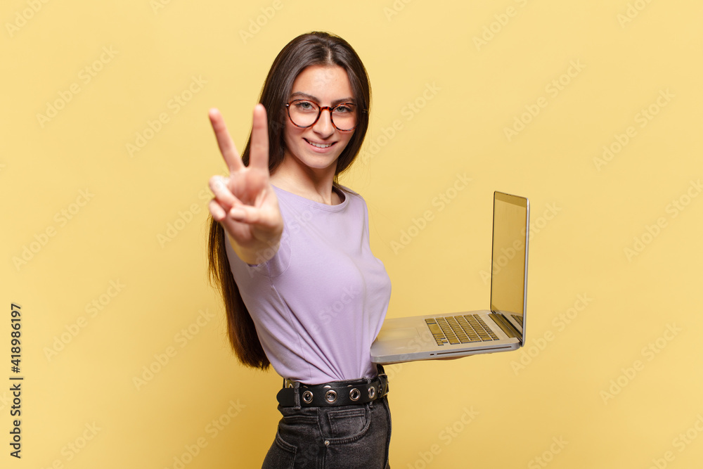 young pretty woman smiling and looking happy, carefree and positive, gesturing victory or peace with one hand. laptop concept - obrazy, fototapety, plakaty 