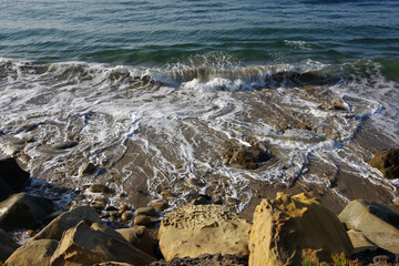 Wall Mural - Water’s edge of the Pacific ocean with waves, sand, surf, rocks, and sun on a warm autumn day in southern California 