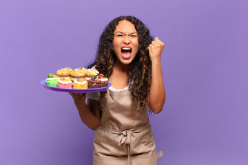 young hispanic woman shouting aggressively with an angry expression or with fists clenched celebrating success. cooking cakes concept