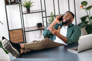 Wall Mural - Relaxed confident african american male freelancer or business man sitting at work desk in the office with legs on table and talking on the cellphone with business partner, gesturing hand, smiling