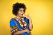© Irene - African american woman wearing african clothing over yellow background covering mouth with hand, shocked and afraid for mistake. surprised expression