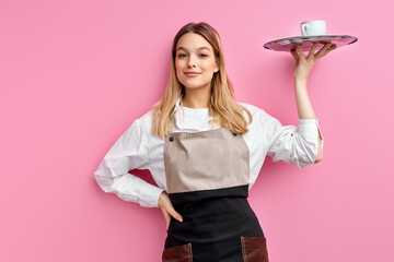 nice woman waitress in apron, offering cup of delicious tasty coffee on tray, stand smiling, friendly staff of restaurant. isolated over pink studio background