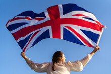The Flag Of England Waving In The Hands Of A Woman Against The Blue Sky. View From The Back. Great Britain, Victory And Success