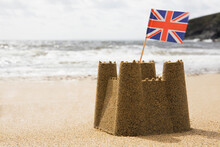 Sandcastle On Empty British Beach With UK Union Jack Flag Flying