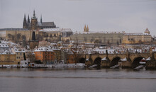 Touristic View Of Beautiful Snowing Prague, Vltava River, Charles Bridge And Prague Castle. The Red Roofs Of The Houses Are Covered With Snow. 