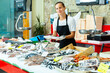 Female fishmonger standing behind seafood shop counter, offering fresh sea bass for sale
