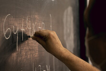 Teacher Wearing Mask Writing Equations On A Blackboard. Covid Situation, Pandemic, New Normal. Close-up, Perspective From The Side.