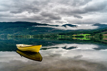 A Single Yellow Row Boat Floats In A Perfectly Calm Lake Which Creates A Beautiful Reflection Of The Low Clouds And Landscape Background