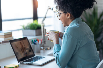 Wall Mural - Beautiful young afro business woman making video call with laptop while drinking coffee sitting in the office at home.