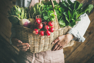 Woman farmer in linen apron holding basket of fresh organic garden vegetables and greens in hands, rustic wooden wall at background