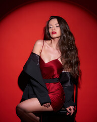 Young beautiful sexy woman with make-up and wavy brunette hair in red silk dress posing against red background in circle of light (spot gobo light)