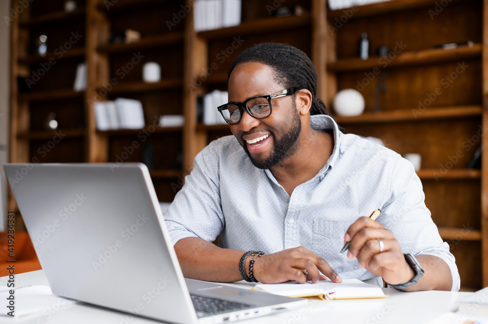 Smiling African-American guy takes notes watching webinars on the laptop,researching business tasks, a male student is studying online, listening video lectures, writes down, e-learning concept - obrazy, fototapety, plakaty 